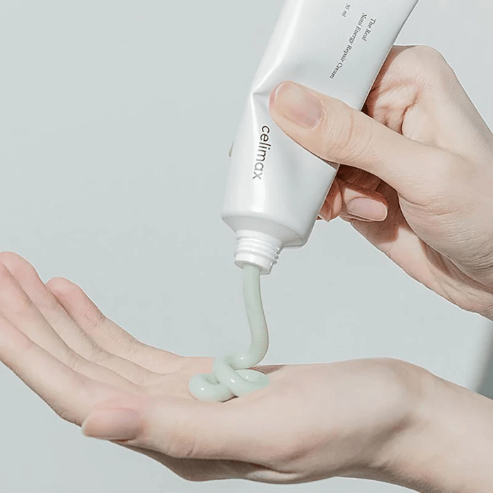 En hand håller en vit tub med Celimax Noni Energy Repair Cream, och krämen pressas ut i en jämn stråle på handflatan. Krämen är ljusgrön och marknadsförs för att ge näring och återställa huden.
