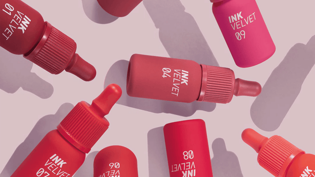 Bilden visar en samling av Peripera Ink The Velvet läppfärger i olika röda och rosa nyanser, med deras karaktäristiska dropp-applikatorer. Dessa produkter är kända för sin intensiva pigmentering och långvariga, sammetsmjuka finish.