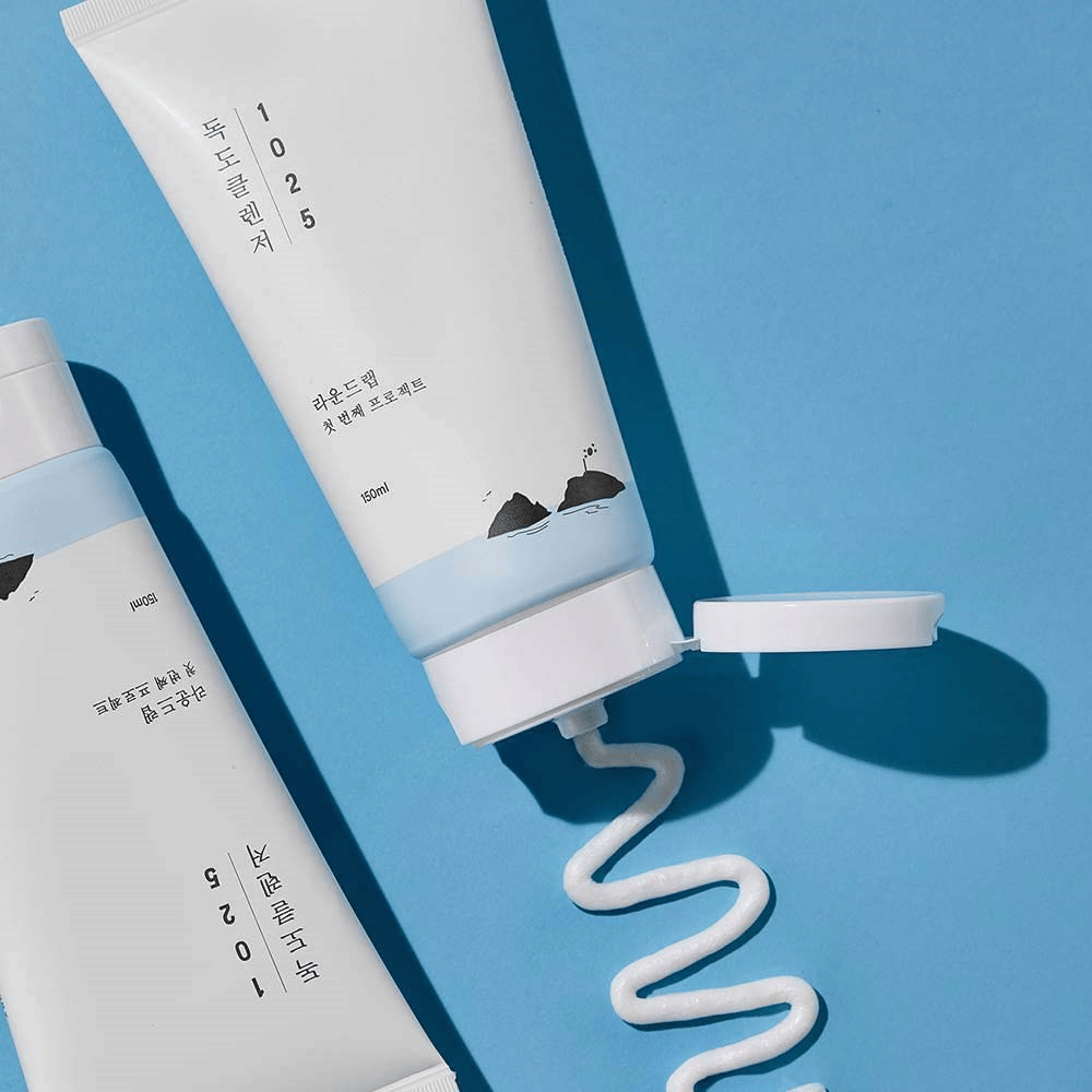 Dwie tuby 1025 Dokdo Cleanser na niebieskim tle, jedna z otwartym korkiem i falistym paskiem wyciśniętego środka czyszczącego.