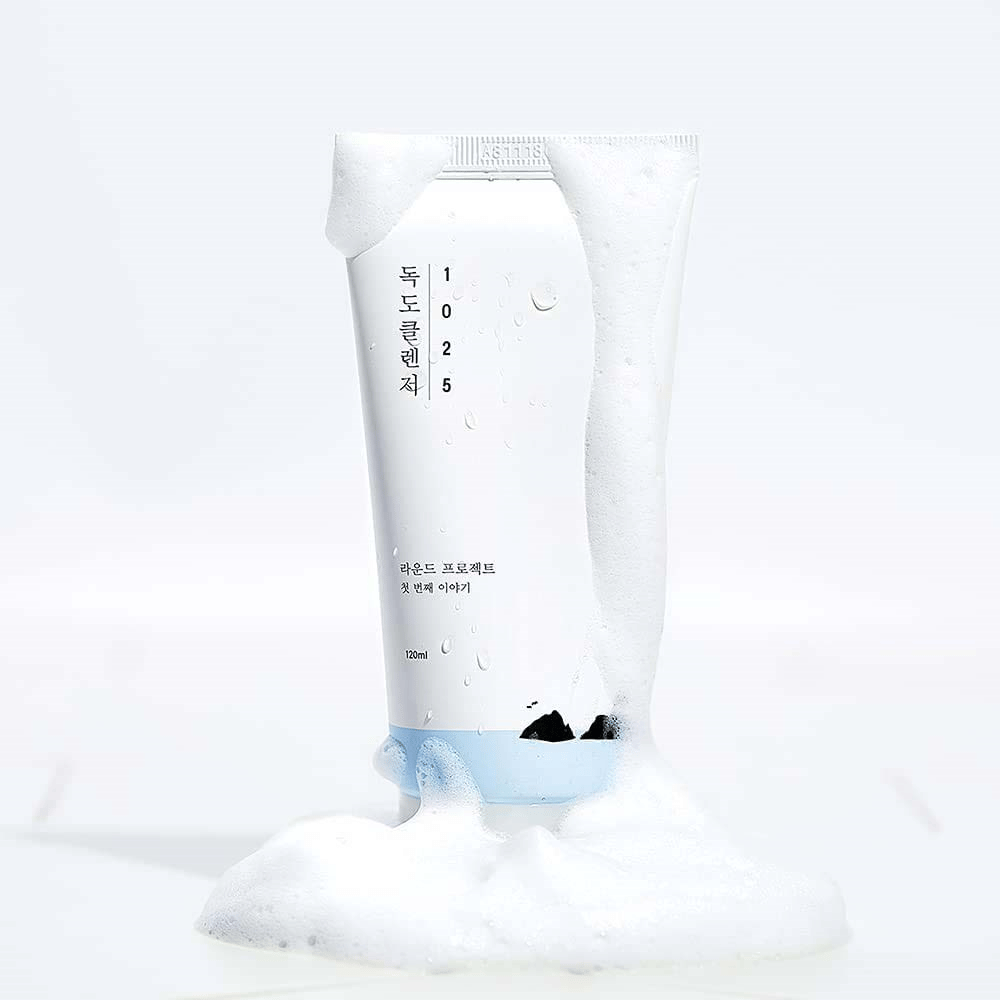 1025 Dokdo Cleanser w białej tubie pokrytej pianą, z kroplami wody i pianą wokół podstawy, wycięta na jasnym tle.