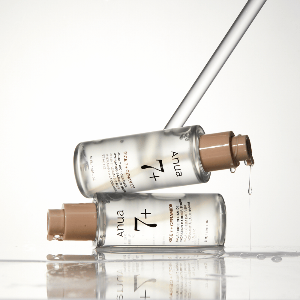 Zdjęcie przedstawia dwie przezroczyste butelki "Anua 77 Rice + Ceramide Hydrating Barrier Serum" z kroplami spływającymi z pipety. Serum, wzbogacone ryżem i ceramidami, jest przeznaczone do nawilżania i wzmacniania bariery skórnej. Butelki są elegancko zaprojektowane, co podkreśla poczucie premium produktu.