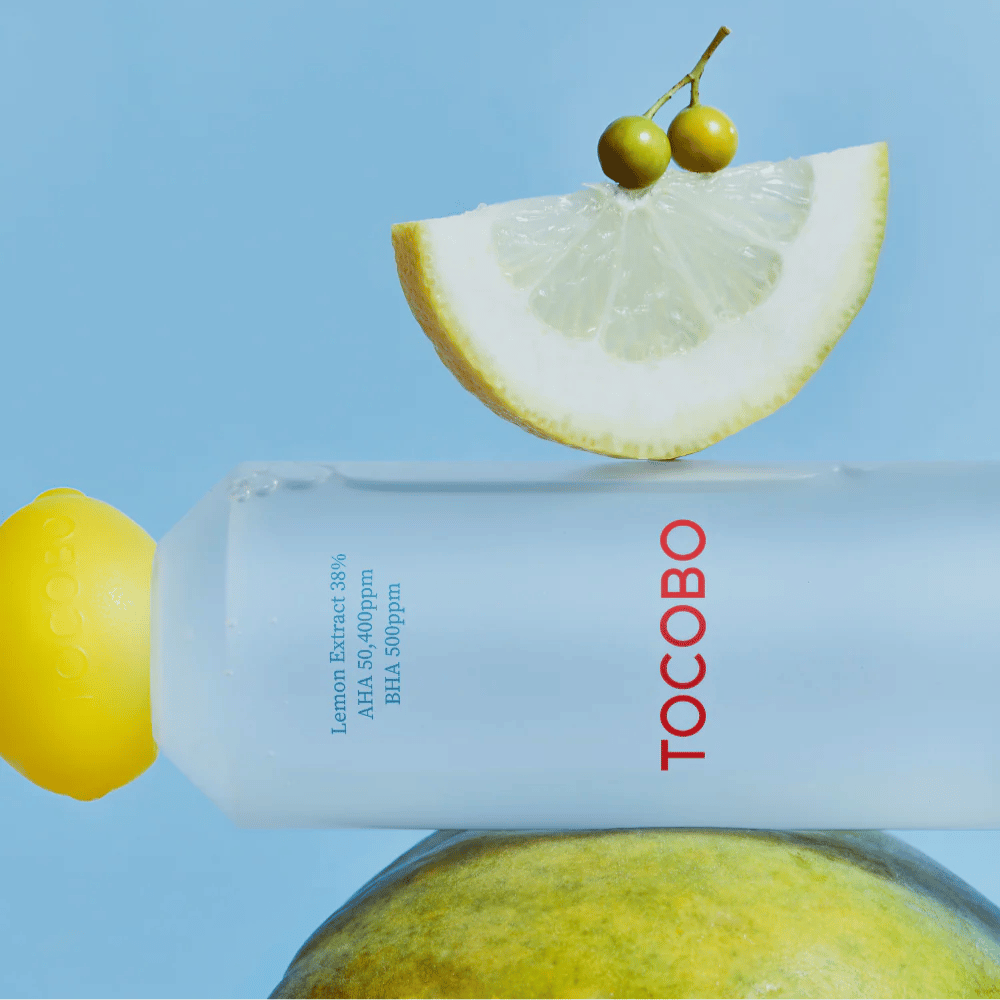 Butelka TOCOBO AHA BHA Lemon Toner, ozdobiona nakrętką w kształcie cytryny. Plasterek cytryny i małe zielone jagody balansują na butelce na niebieskim tle.