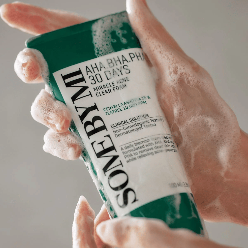 Osoba trzyma tubę "SOME BY MI AHA, BHA, PHA 30 Days Miracle Acne Clear Foam" z pieniącą się zawartością na dłoniach. Produkt przeznaczony jest do codziennego użytku, aby zwalczać trądzik i oczyszczać pory dzięki aktywnym składnikom, takim jak centella, olej tea tree i kwasy AHA, BHA oraz PHA.