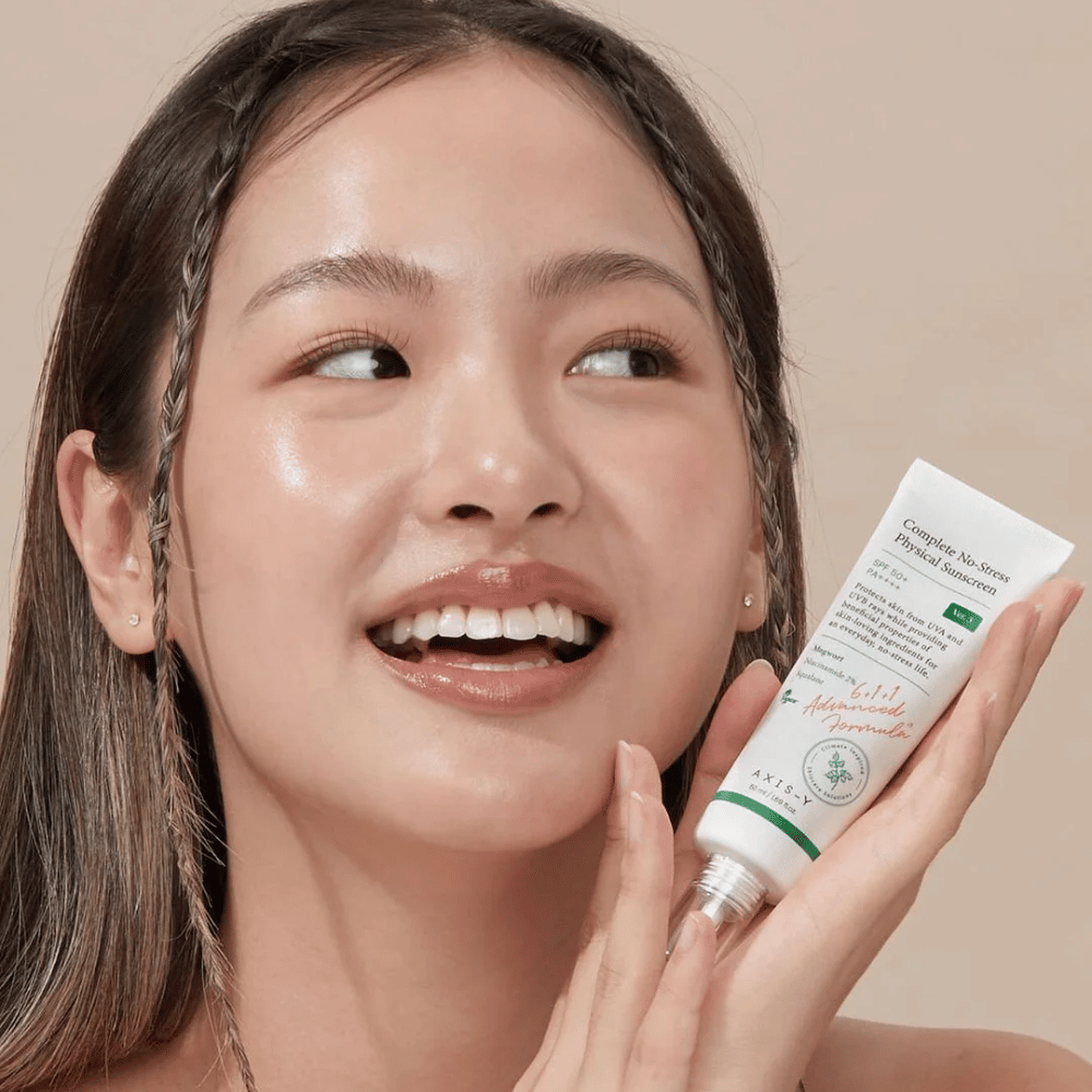 Na zdjęciu widać młodą kobietę, która radośnie się uśmiecha i trzyma tubkę z AXIS-Y Complete No-Stress Physical Sunscreen. Ma promienny i naturalny wygląd z minimalnym makijażem, co podkreśla produkt, który prezentuje.