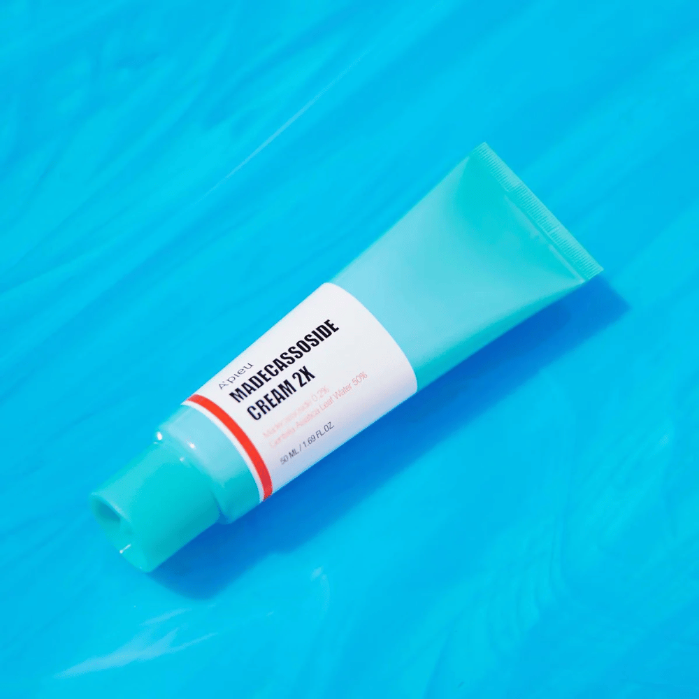 Tubka A'PIEU Madecassoside Cream 2X leży ukośnie na niebieskim tle z zmarszczkami przypominającymi wodę. Tubka ma białe ciało z jasnoturkusową nakrętką i czerwoną do pomarańczowej etykietę, na której wyraźnie napisano nazwę produktu i aktywny składnik Madecassoside 2%. Na etykiecie znajduje się również ilość, która wynosi 50 ml / 1.8 fl oz. Jasna i kolorowa prezentacja daje świeże i energetyczne wrażenie.