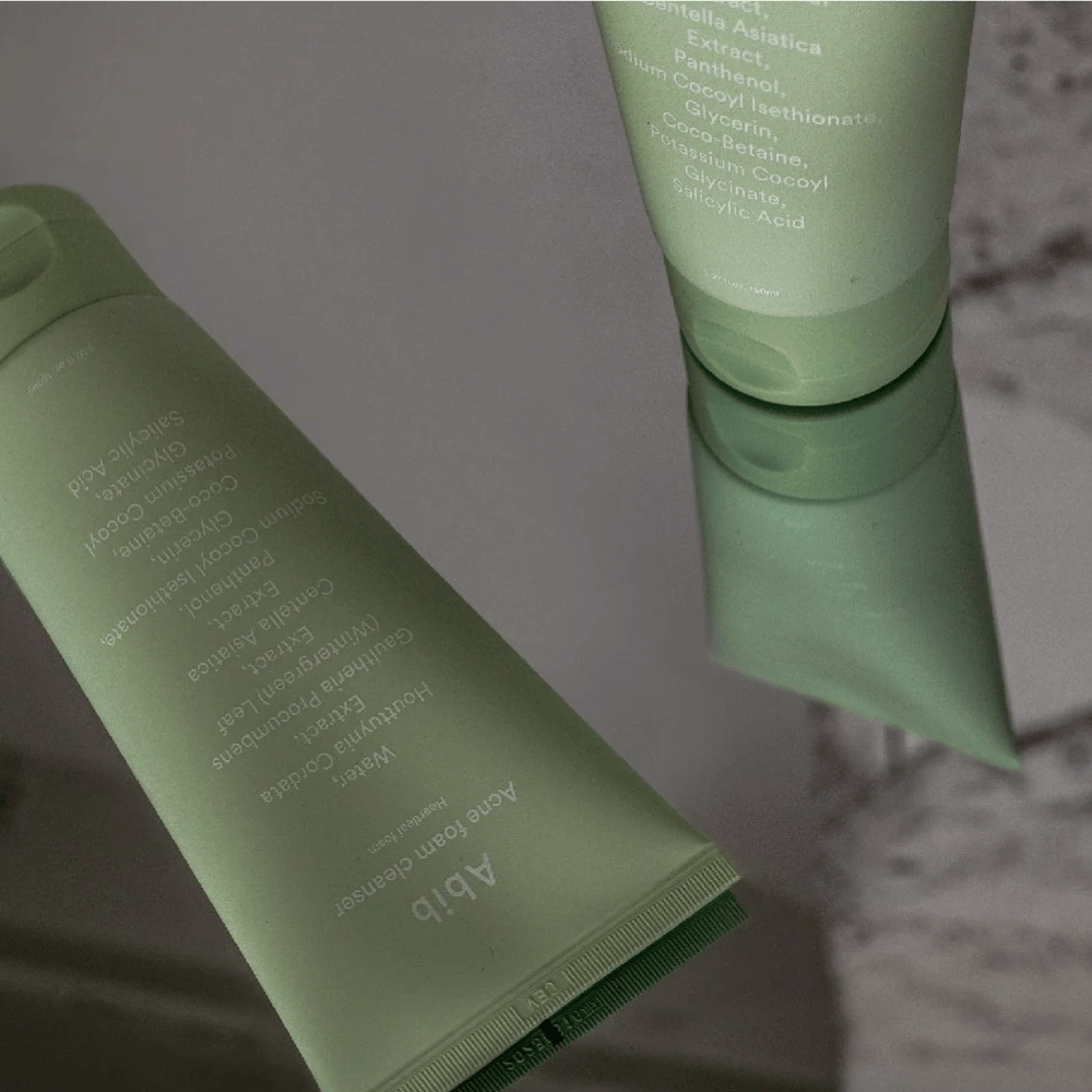 Zielona tubka Abib Acne Foam Cleanser Heartleaf leżąca na lustrzanym stole z przodem produktu i tekstem odbitym w lustrze. Składniki takie jak kwas salicylowy, centella i ekstrakt z liści serca są wyraźnie widoczne na opakowaniu.