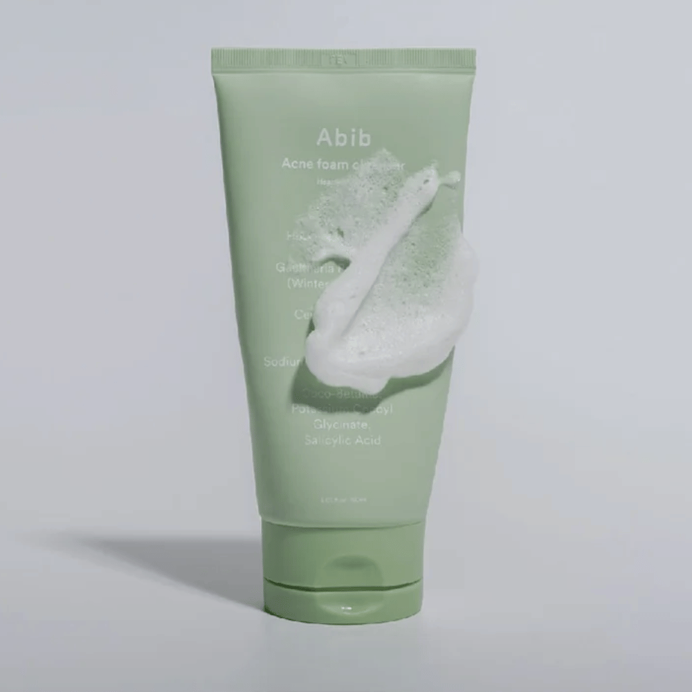 Zielona tubka z Abib Acne Foam Cleanser Heartleaf, pokryta białą pianą ilustrującą efekt pieniącego się oczyszczania. Produkt zawiera ekstrakt z liści serca i kwas salicylowy do skóry skłonnej do trądziku.
