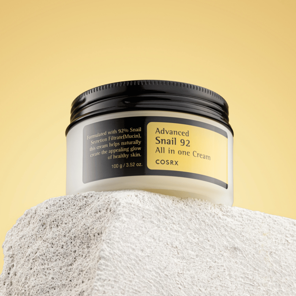 Słoik COSRX Advanced Snail 92 All in one Cream umieszczony na kamienistej powierzchni z żółtym tłem.