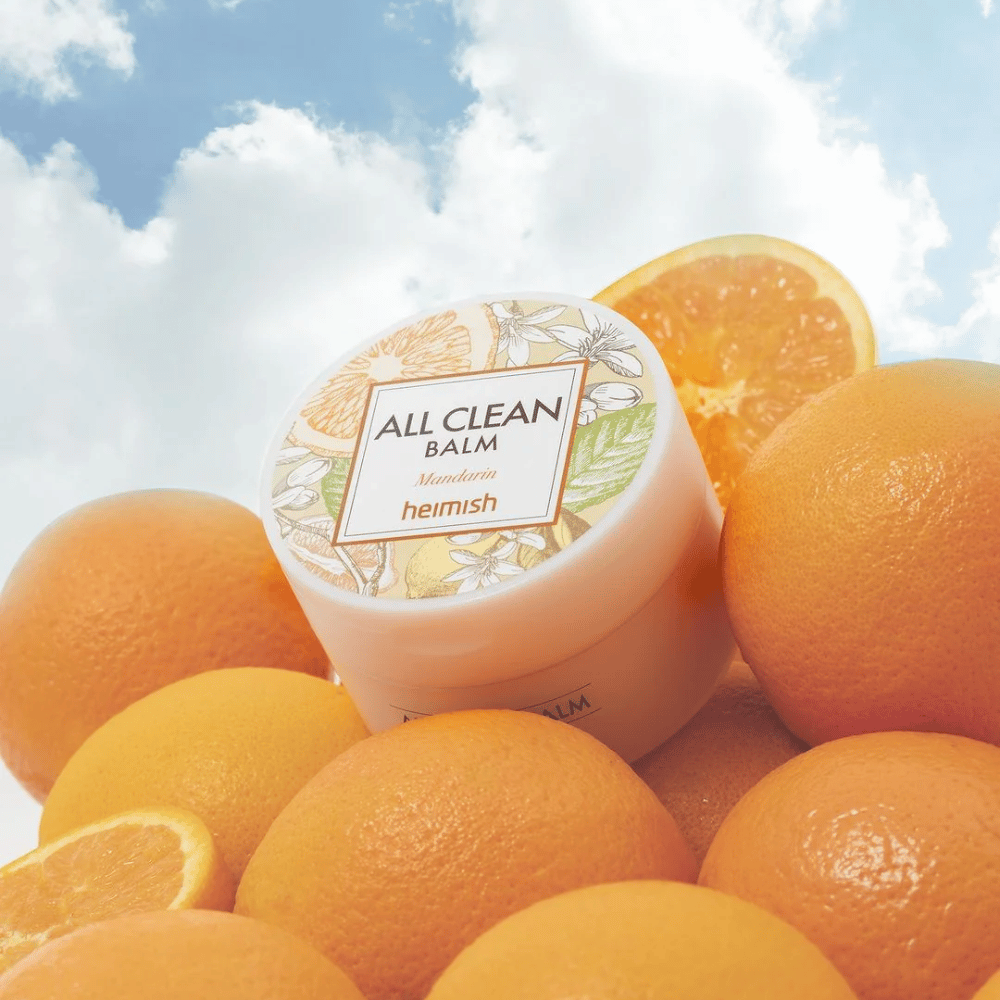 Słoik Heimish All Clean Balm Mandarin umieszczony na stosie pomarańczy, z niebieskim niebem w tle.