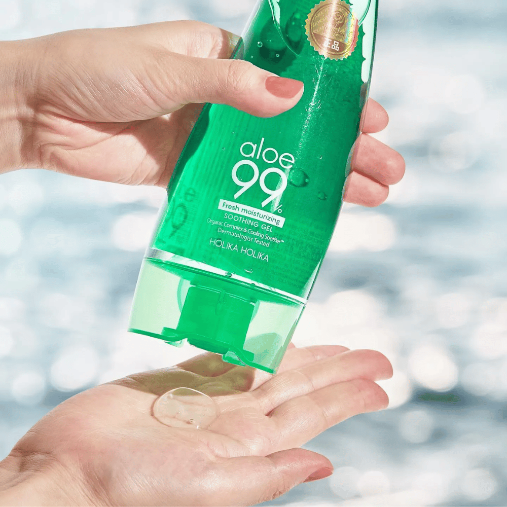 Ręka trzyma zieloną tubkę z Holika Holika Aloe 99% Soothing Gel, podczas gdy druga ręka łapie porcję żelu, który jest wydawany. Tło jest rozmyte i pokazuje wodnisty, błyszczący wzór.