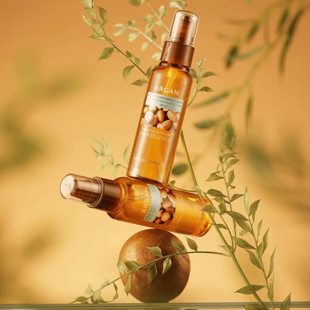 Dwie butelki Nature Republic Argan Essential Moisture Hair Oil umieszczone wśród zielonych liści – jedwabisty połysk dla suchych włosów.