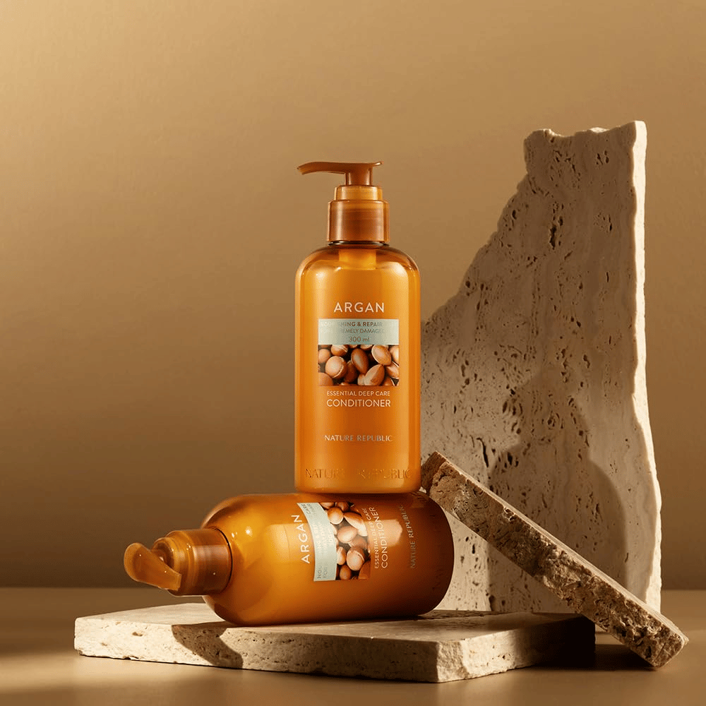 Dwie butelki Nature Republic Argan Essential Moisture Repairing Conditioner na kamiennej powierzchni w ciepłym tonie.
