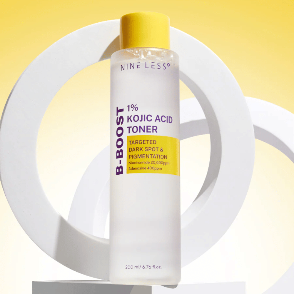NINE LESS B-Boost 1% Kojic Acid Toner stojący na żółtym tle z napisem "Targeted Dark Spot & Pigmentation". Tonik wyrównujący koloryt skóry z niacynamidem i adeniną.
