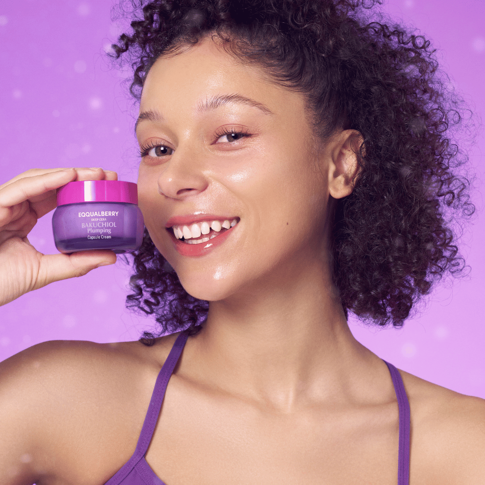Kobieta z promienną cerą trzyma EQQUALBERRY Bakuchiol Plumping Capsule Cream i uśmiecha się na tle fioletowego tła.