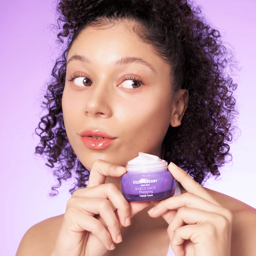 Kobieta trzyma EQQUALBERRY Bakuchiol Plumping Capsule Cream w ręce i prezentuje swoją nawilżoną, promienną skórę na tle fioletowego tła.