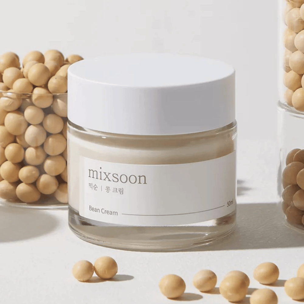 Przezroczysty słoik z białą etykietą i pokrywką, oznaczony "mixsoon 팥 크림" oraz "Bean Cream" z podaną objętością "50ml". Słoik zawiera jasnożółty krem i jest umieszczony na jasnej powierzchni. Obok słoika znajduje się szklany pojemnik pełen jasnobrązowych ziaren, które rozsypały się na powierzchni, co sugeruje, że krem zawiera ekstrakt z ziaren. Tło i oświetlenie tworzą ciepłą i naturalną atmosferę.