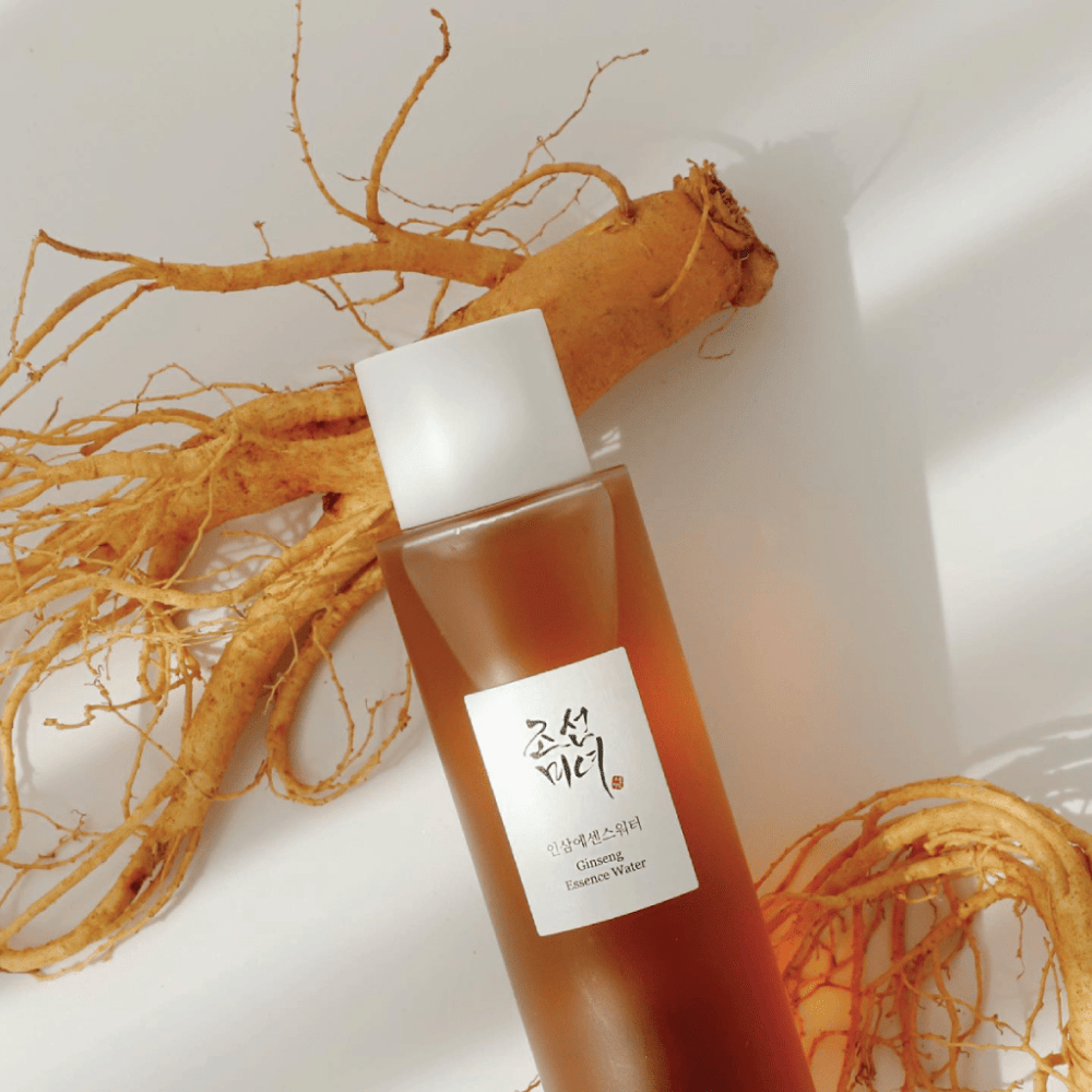 Brązowa butelka Beauty of Joseon Ginseng Essence Water z białą nakrętką leży obok świeżego korzenia żeń-szenia na białym tle.