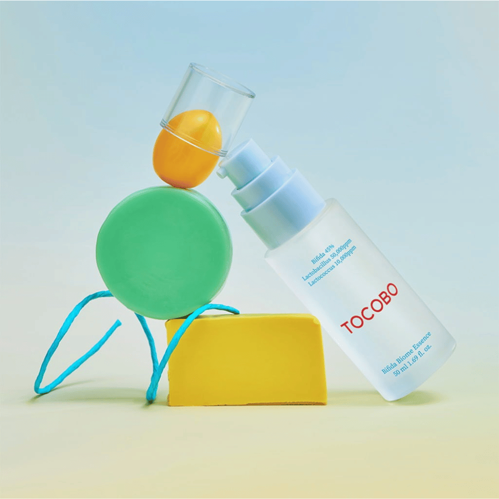 TOCOBO Bifida Biome Essence w matowej butelce z pompką z czerwoną logotypą, umieszczoną na kolorowym tle z zielonymi, żółtymi i pomarańczowymi geometrycznymi kształtami. Tekst na butelce wskazuje Bifida 5%, Lactobacillus 3 000 ppm i Lactococcus 1 000 ppm.