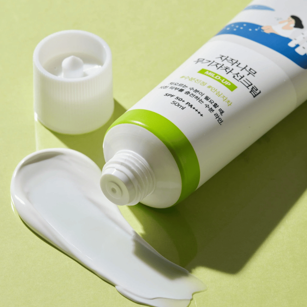 Zbliżenie na Birch Juice Moisturizing Mild Up Sunscreen z odkręconą nakrętką, biały krem przeciwsłoneczny rozmazany na jasnym zielonym tle, SPF 50+ PA++++, 50 ml.