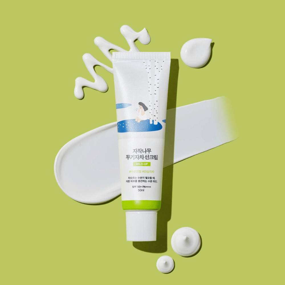 Birch Juice Moisturizing Mild Up Sunscreen – biała tubka z zieloną bazą na zielonym tle, otoczona rozmazanym kremem przeciwsłonecznym i kroplami, SPF 50+ PA++++, 50 ml.