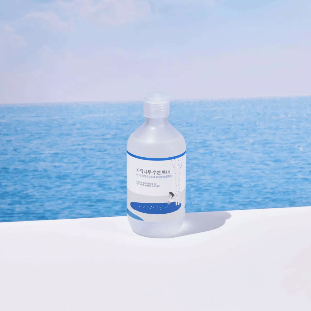 Butelka ROUND LAB Birch Juice Moisturizing Toner stoi na białej powierzchni, z morzem i błękitnym niebem w tle