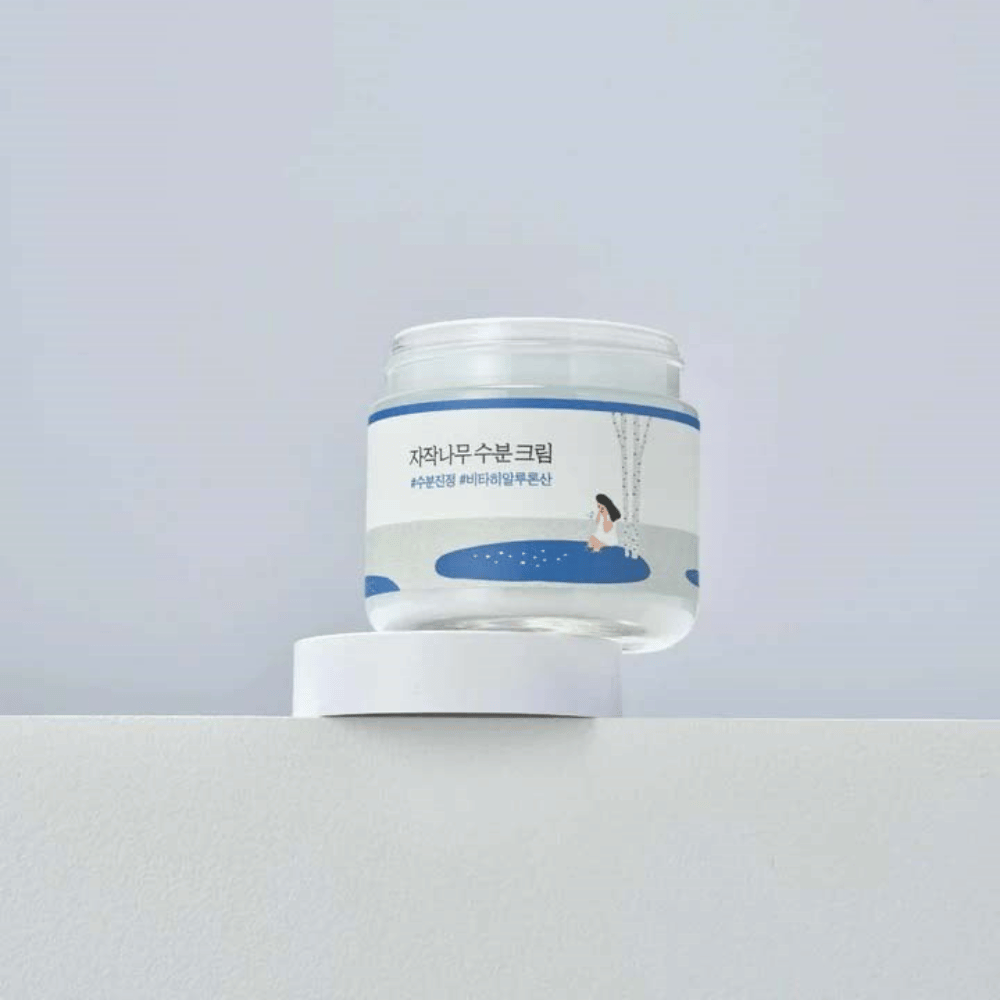 Round Lab Birch Juice Moisturizing Cream w przezroczystym słoiku z białą etykietą i niebieską ilustracją kobiety przy brzozie, stojącym na białej powierzchni na jasnym tle.