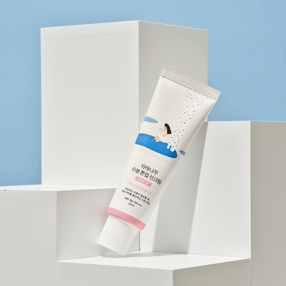 Tuba z Birch Juice Moisturizing Tone Up Sunscreen umieszczona na białych kostkach na jasnoniebieskim tle. Tuba jest biała z ilustracją kobiety przy brzozie i wodzie. Tekst po koreańsku wskazuje, że produkt ma SPF 50+ PA++++ i zawiera 50 ml.
