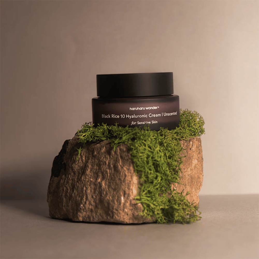 Zdjęcie przedstawia słoik kremu do skóry oznaczony "haruharu wonder Black Rice 10 Hyaluronic Cream Unscented for Sensitive Skin". Słoik jest czarny i stoi na dużym kamieniu pokrytym jasnozielonym mchem, co tworzy naturalne i przyziemne wrażenie.