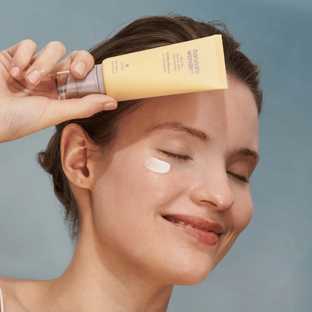 Uśmiechnięta kobieta nakłada Black Rice Moisture Airyfit Daily Sunscreen SPF 50+ PA++++ od Haruharu Wonder na swoją twarz. Trzyma żółtą tubkę z kremem przeciwsłonecznym i ma odrobinę produktu na policzku. Tło jest jasnoniebieskie.