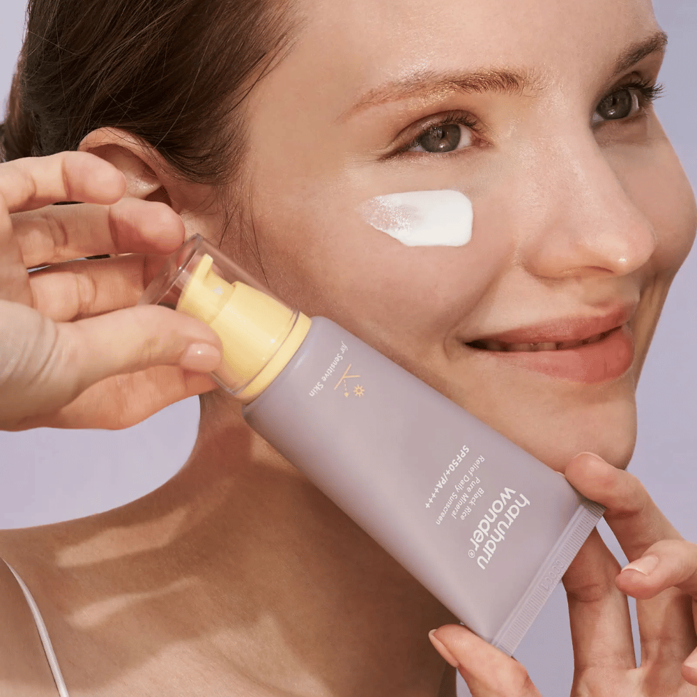 Uśmiechnięta kobieta nakłada Haruharu Wonder Black Rice Pure Mineral Relief Daily Sunscreen SPF 50+ PA++++ na swoją twarz. Trzyma fioletową tubkę z kremem przeciwsłonecznym i ma kropkę produktu na policzku. Tło jest jasnofioletowe.