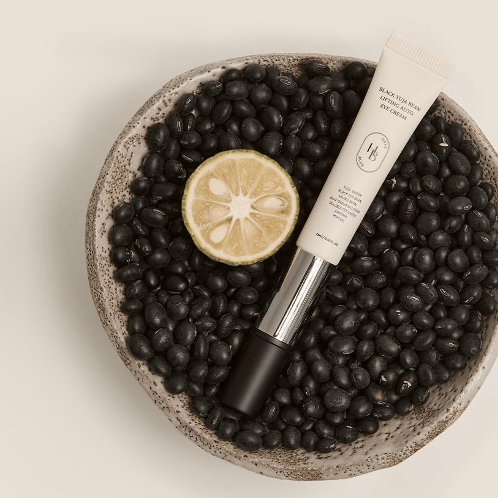 Black Yuja Bean Lifting Auto Eye Cream leżące w misce wypełnionej czarnymi fasolami i połówką owocu cytrusowego – wizualna reprezentacja naturalnych składników produktu.