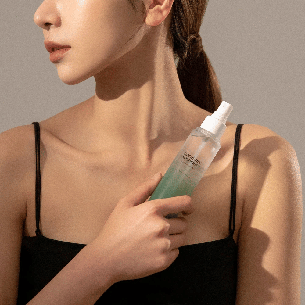 Obraz HaruHaru Wonder Black Bamboo Mist trzymany przez kobietę. Produkt znajduje się w przezroczystej butelce z rozpylaczem o zielonym odcieniu i białej pompce. Kobieta ma swobodny wygląd, nosi czarną bluzkę i skupia się na używaniu mgiełki, aby zapewnić skórze nawilżenie i blask. Butelka jest częścią rutyny pielęgnacyjnej skóry.