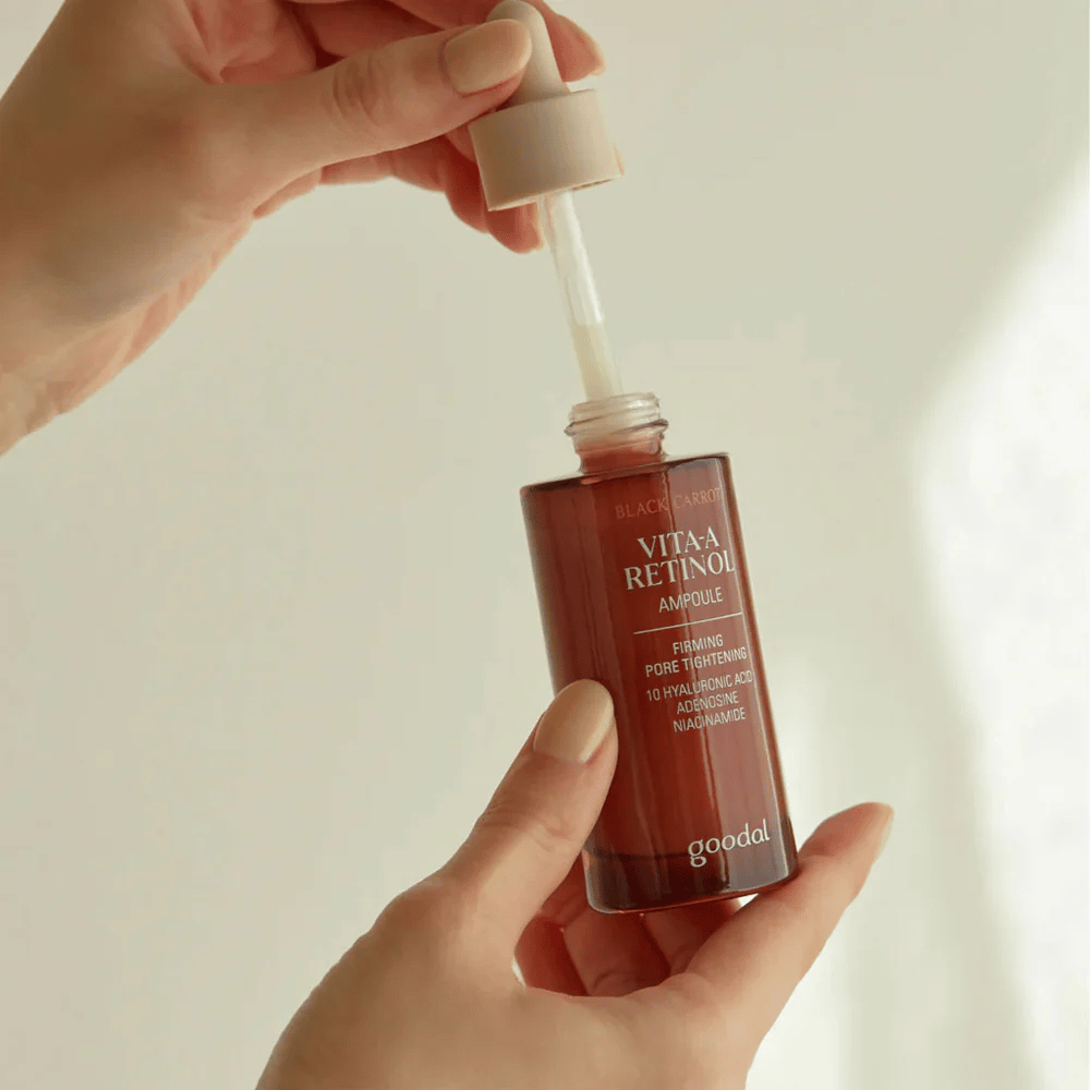 Ręka trzyma butelkę z Goodal Black Carrot Vita-A Retinol Firming Ampoule. Etykieta butelki pokazuje nazwę produktu i właściwości, w tym "Ujędrniające Zwężanie Porów" oraz składniki takie jak 10% kwasu hialuronowego, adenozynę i niacynamid. Pipeta jest w kolorze beżowym.