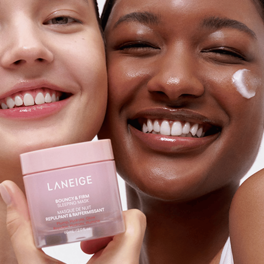 Dwie osoby uśmiechają się i trzymają Laneige Bouncy & Firm Sleeping Mask. Jedna z nich nałożyła maskę na swój policzek, a produkt jest wyraźnie widoczny w ich rękach. Maska znajduje się w różowym słoiku i jest reklamowana jako "Plump, Hydrate, Glow."