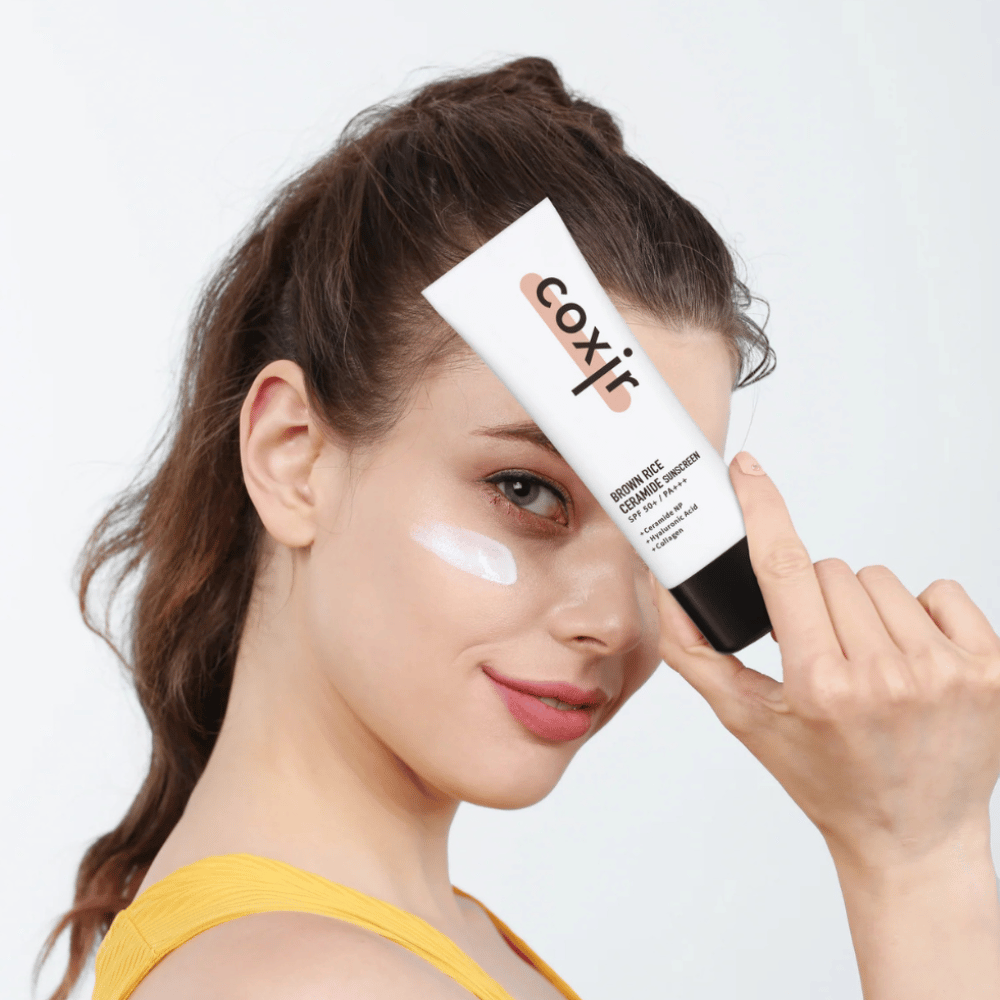 Kobieta z kremem przeciwsłonecznym nałożonym na policzek trzyma tubkę Coxir Brown Rice Ceramide Sunscreen, SPF 50+ PA+++, przy czole i lekko się uśmiecha.