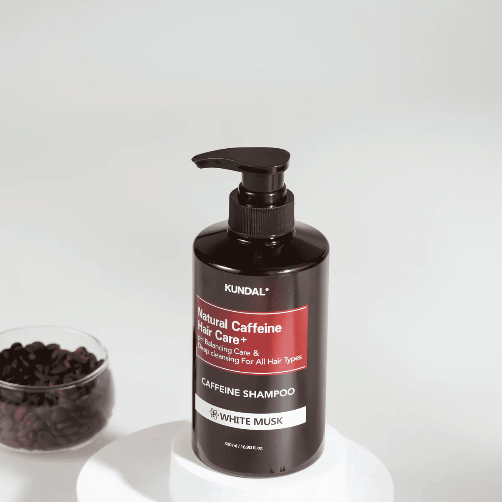 KUNDAL Natural Caffeine Hair Care+ Szampon White Musk w czarnej butelce z pompką, 500 ml, umieszczony obok miski z ziarnami kawy.