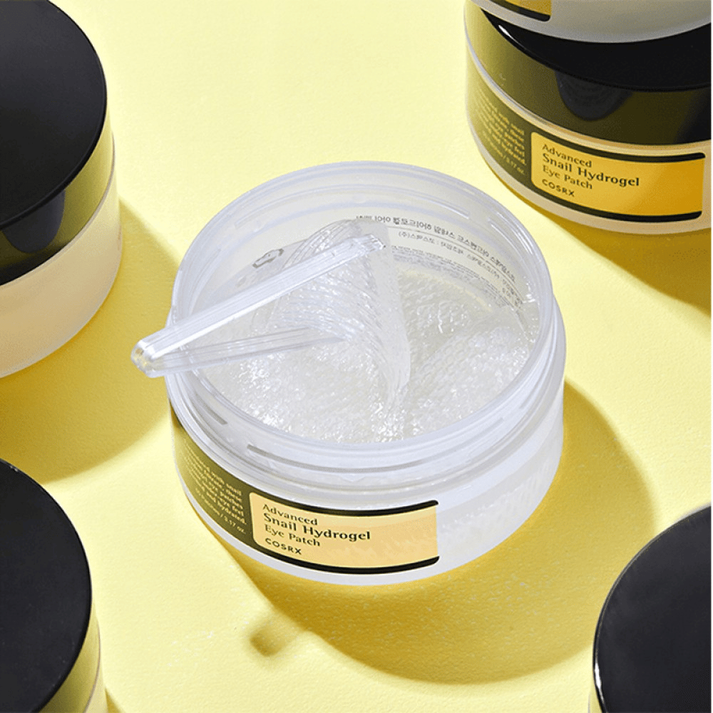 Otwarty słoik COSRX Advanced Snail Hydrogel Eye Patch z przezroczystymi płatkami pod oczy i małą szpatułką, otoczony przez więcej zamkniętych słoików na żółtym tle.