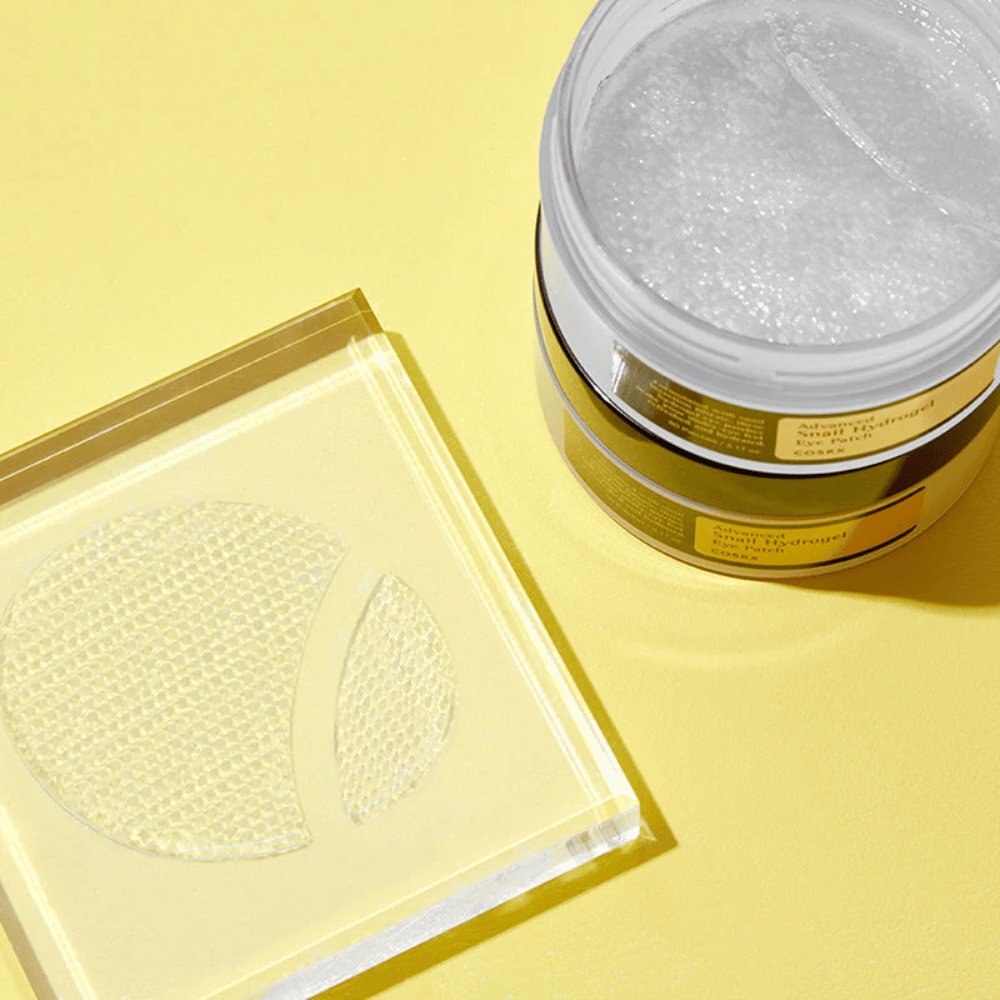 Otwarty słoik COSRX Advanced Snail Hydrogel Eye Patch stoi na żółtym tle obok przezroczystej podstawki, na której leżą dwie płatki pod oczy.