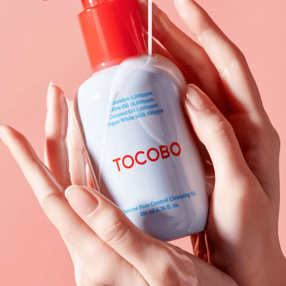 TOCOBO Calamine Pore Control Cleansing Oil trzymane w rękach na różowym tle, czerwona pompka butelki i lista składników są wyraźnie widoczne.