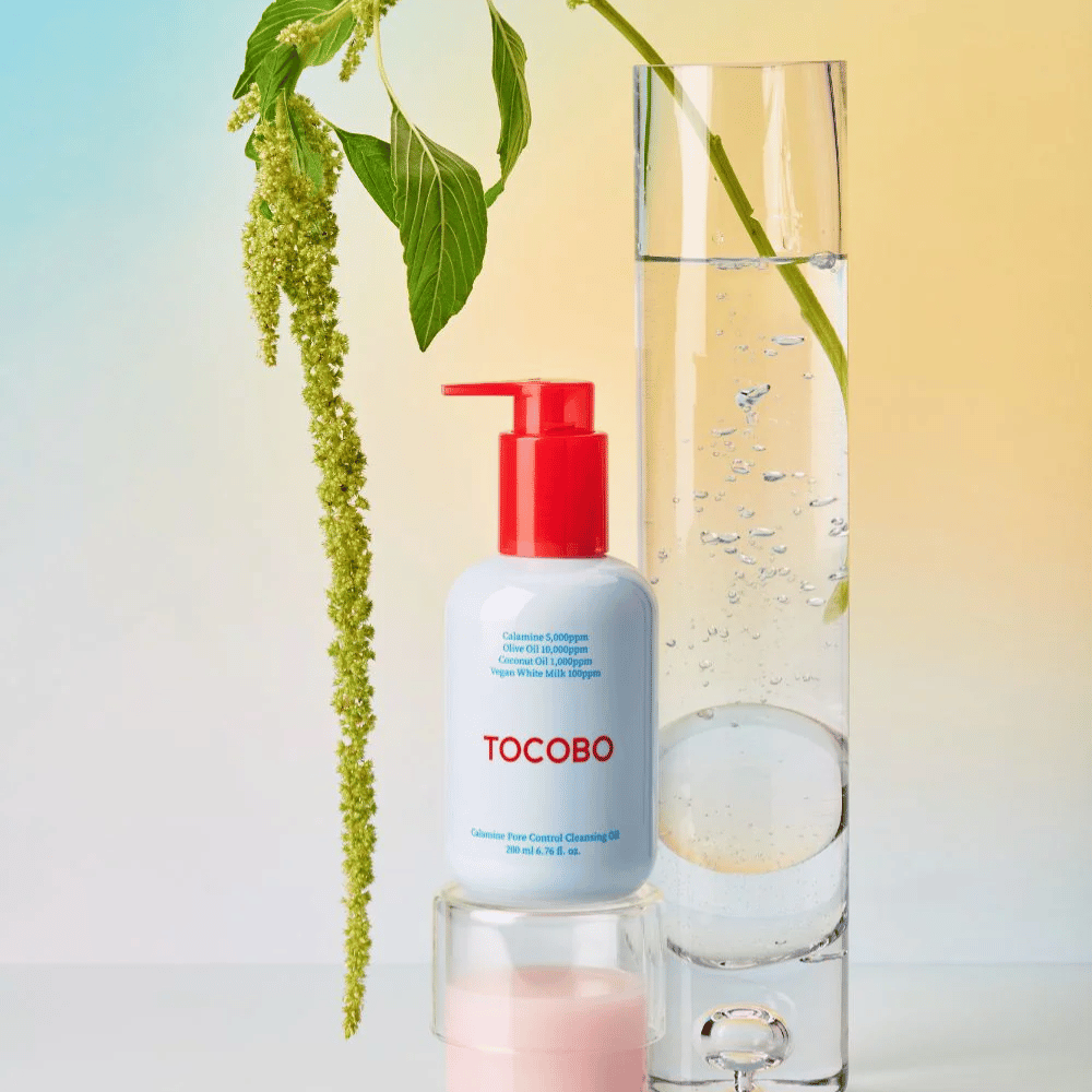 TOCOBO Calamine Pore Control Cleansing Oil z czerwoną pompką stoi obok wysokiej szklanej probówki wypełnionej wodą i zieloną rośliną, na jasnym pastelowym tle.