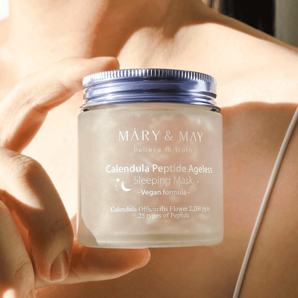 Kobieta trzyma słoik z MARY & MAY Calendula Peptide Ageless Sleeping Mask. Słoik jest przezroczysty z fioletowym wieczkiem, a napis "Calendula Peptide Ageless Sleeping Mask" jest wyraźnie widoczny. Produkt to wegańska maska na noc z nagietkiem i peptydami, przeznaczona do nadania skórze młodzieńczego blasku w nocy.