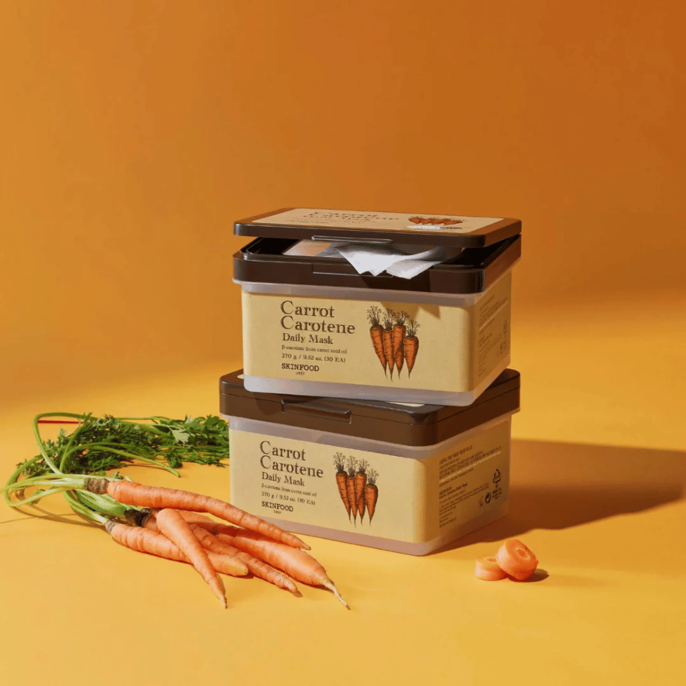 Obraz przedstawia trzy ułożone opakowania "Carrot Carotene Daily Mask" od SKINFOOD, umieszczone na pomarańczowym tle wraz ze świeżymi marchewkami. Każde pudełko zawiera maseczki do twarzy wzbogacone w β-karoten z oleju z nasion marchwi, zaprojektowane do codziennego użytku, aby odżywiać i pielęgnować skórę.