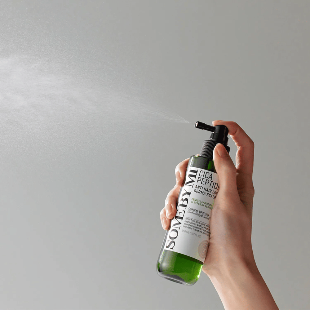 Ręka spryskująca SOME BY MI Cica Peptide Anti Hair Loss Derma Scalp Tonic z zielonej butelki z czarną dyszą i białą etykietą na szarym tle.