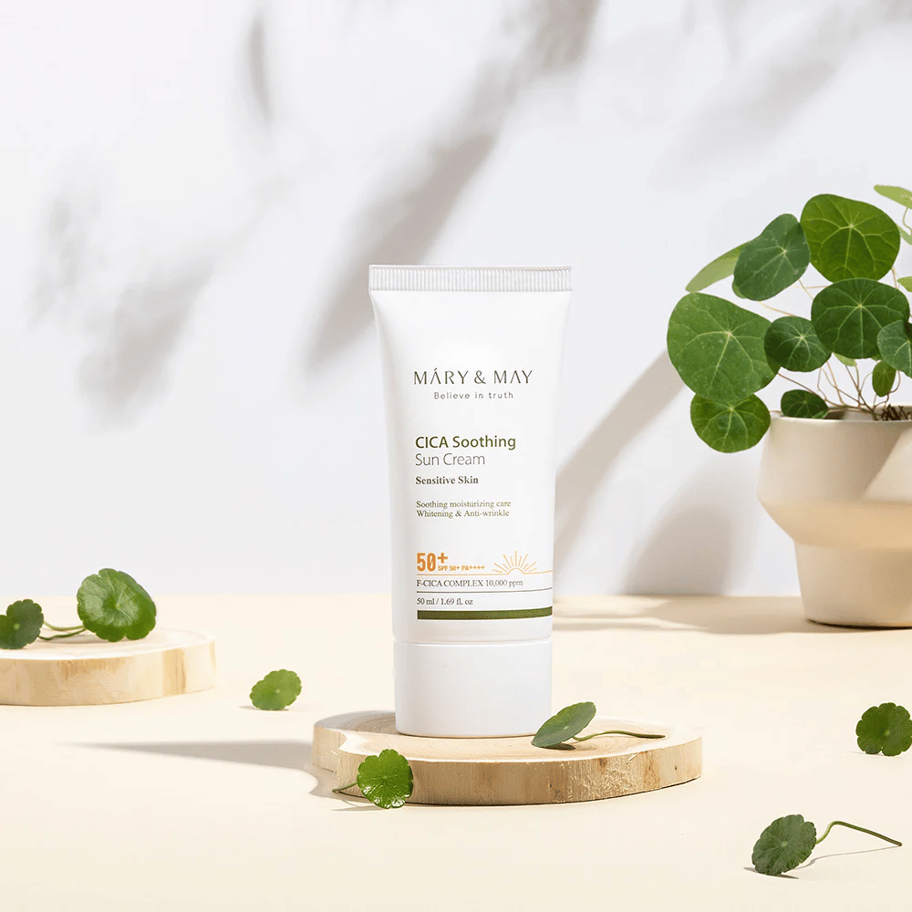 Mary&May CICA Soothing Sun Cream umieszczony na drewnianym podium, otoczony zielonymi liśćmi centelli. Obraz podkreśla składniki łagodzące skórę oraz naturalną inspirację stojącą za produktem.
