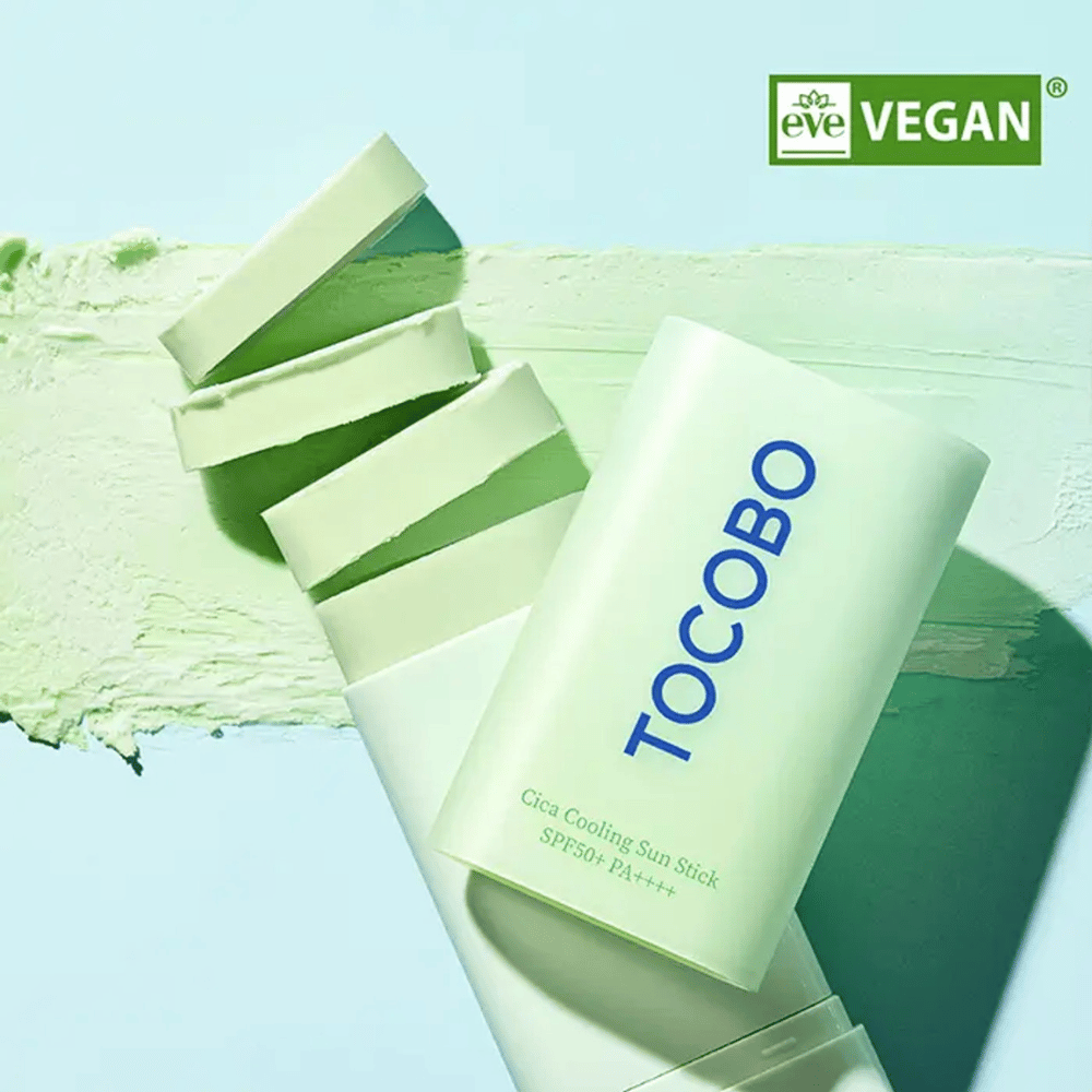 Tocobo Cica Cooling Sun Stick SPF50+ PA++++ w jasnozielonym opakowaniu z oznaczeniem wegańskim (EVE VEGAN), leżącym na warstwie zielonego sztyftu do opalania.