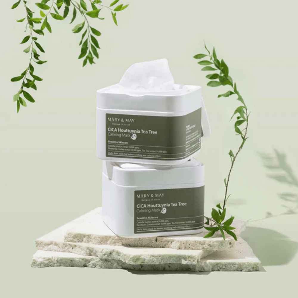 Dwa słoiki Mary & May Cica Houttuynia Tea Tree Calming Mask umieszczone na naturalnym kamieniu z zielonymi detalami roślinnymi – kojące maseczki w płachcie dla skóry wrażliwej.