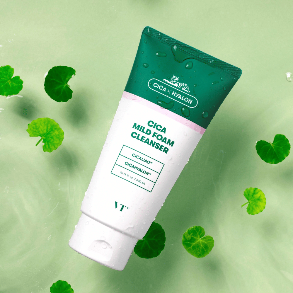 VT Cica Mild Foam Cleanser leży na jasnozielonym podłożu z kroplami wody na opakowaniu i jest otoczony zielonymi liśćmi centella asiatica, co podkreśla kojące właściwości produktu.