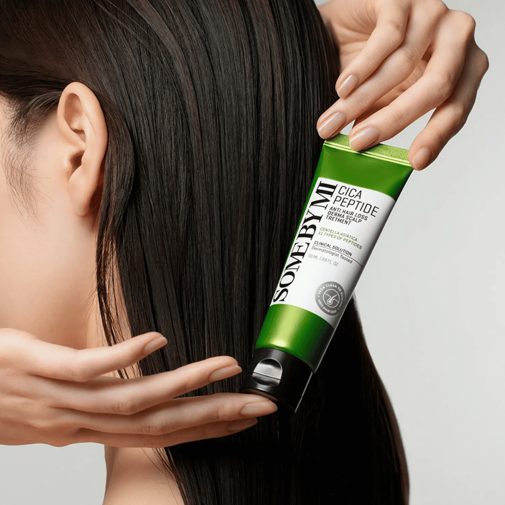 Kobieta nakłada SOME BY MI Cica Peptide Anti Hair Loss Derma Scalp Treatment na długie, ciemne włosy za pomocą zielonej i białej tuby.