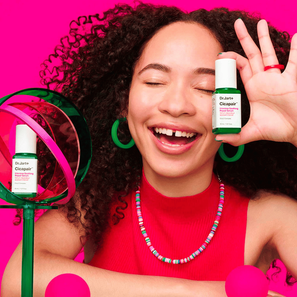 Kobieta szeroko się uśmiecha, trzymając Dr.Jart+ Cicapair Intensive Soothing Repair Serum przed twarzą. Ma na sobie zielone kolczyki i czerwony naszyjnik. Tło jest jasnoróżowe i ozdobione zielonymi i różowymi kółkami.