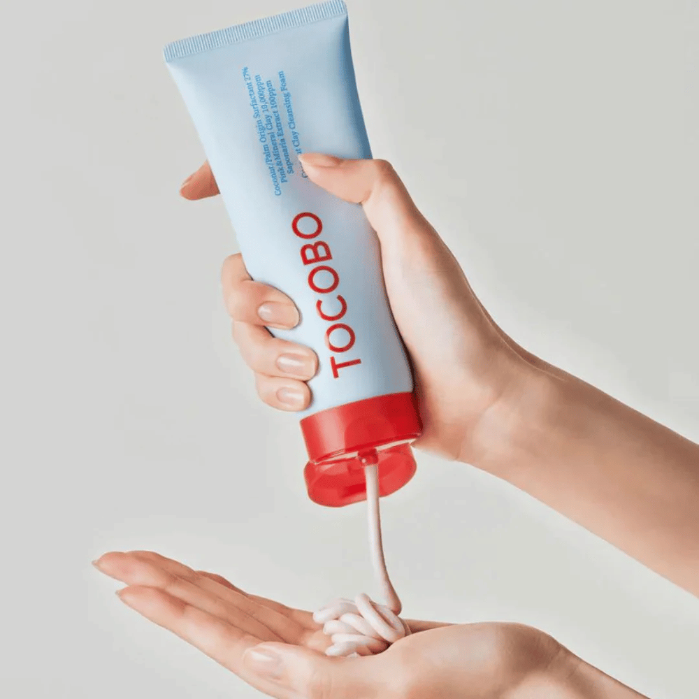 Osoba trzyma tubę TOCOBO Coconut Clay Cleansing Foam. Tuba jest jasnoniebieska z czerwoną nakrętką i napisem "TOCOBO" z przodu. Produkt jest wyciskany z tuby i formowany w ręku jak szampon. Tło jest neutralne, co skupia uwagę na produkcie.